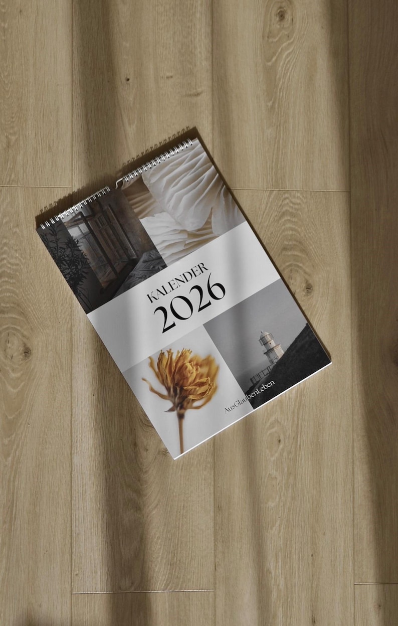 May include: A spiral-bound 2026 calendar with a collage of images, including a flower, architectural details, and a lighthouse. The word "KALENDER" and the year "2026" are prominently displayed. The calendar is lying on a wooden surface.