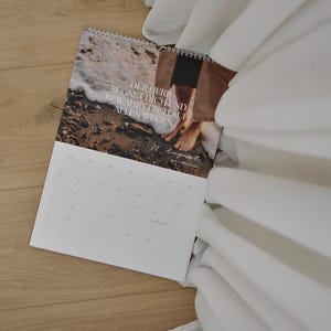 May include: A spiral-bound calendar with a beach scene and the text "DER HERR SEGNET DICH UND BEWAHRT DICH AUF ALLEN WEGEN." The calendar is open to August, resting on a wooden surface, with white fabric in the background.