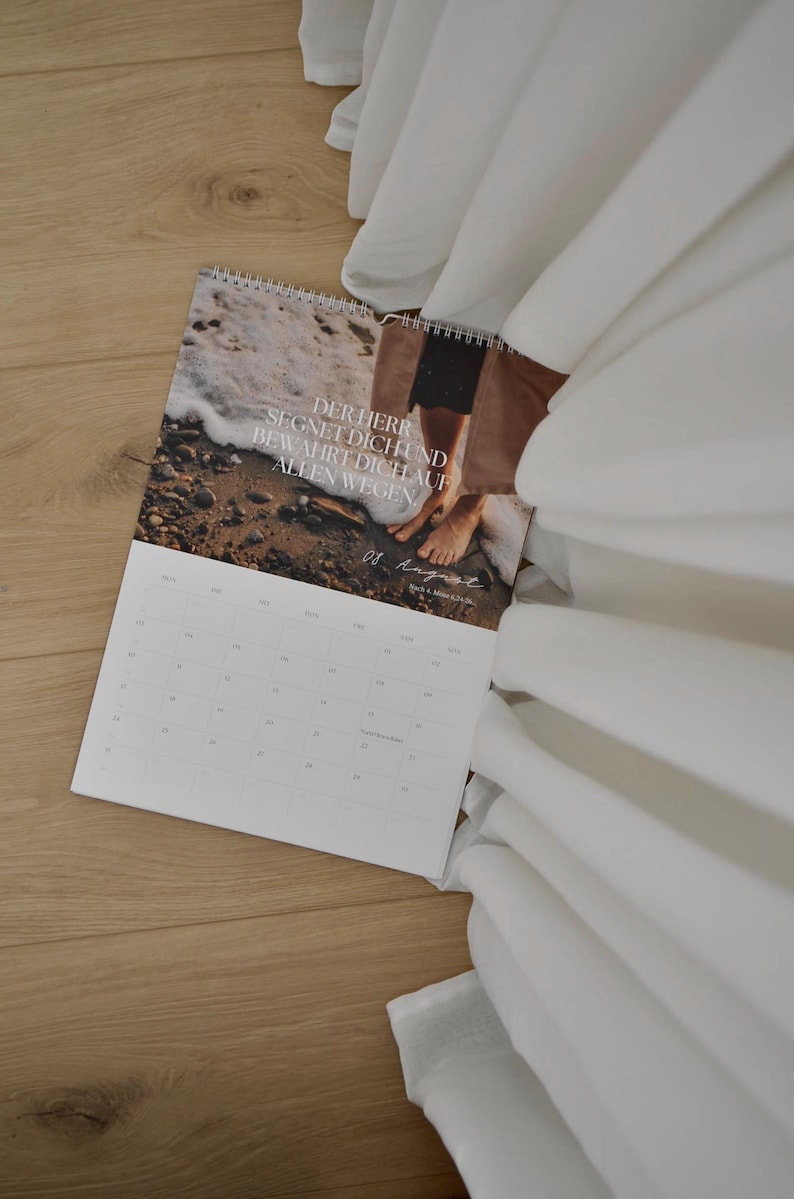 May include: A spiral-bound calendar with a beach scene and the text "DER HERR SEGNET DICH UND BEWAHRT DICH AUF ALLEN WEGEN." The calendar is open to August, resting on a wooden surface, with white fabric in the background.