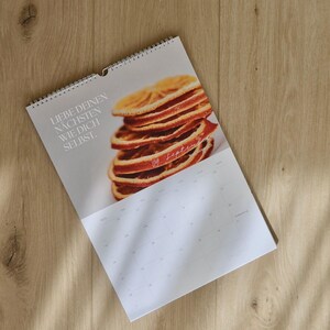 May include: A spiral-bound calendar featuring a stack of dried orange slices. The calendar has the text "LIEBE DEINEN N&Auml;CHSTEN WIE DICH SELBST." and the date "09 September" printed on the page. The calendar is on a wooden surface.