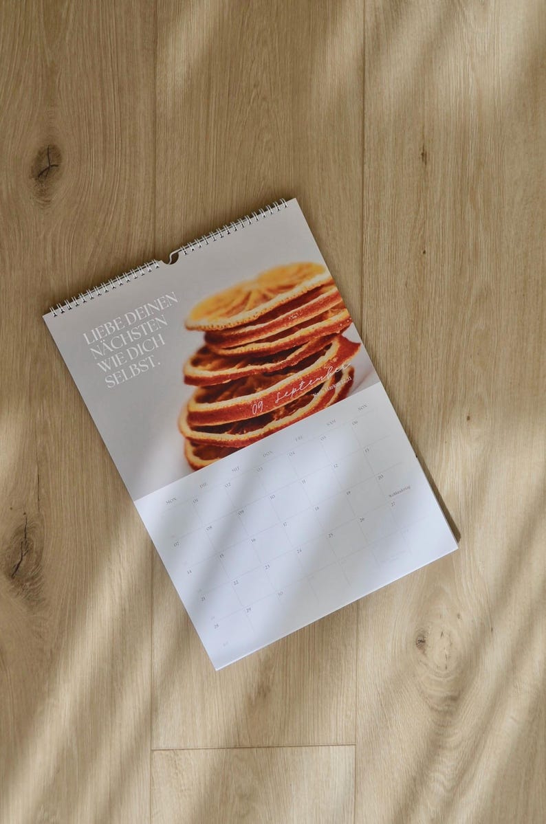 May include: A spiral-bound calendar featuring a stack of dried orange slices. The calendar has the text "LIEBE DEINEN N&Auml;CHSTEN WIE DICH SELBST." and the date "09 September" printed on the page. The calendar is on a wooden surface.
