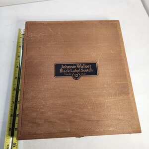 Puede incluir: Una caja de madera marr&oacute;n con una etiqueta azul que dice "Johnnie Walker Black Label Scotch 12 Years Old". La caja es rectangular. Una regla est&aacute; colocada en el lado izquierdo.