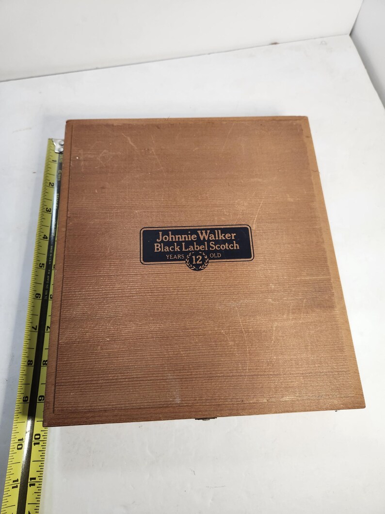 Puede incluir: Una caja de madera marr&oacute;n con una etiqueta azul que dice "Johnnie Walker Black Label Scotch 12 Years Old". La caja es rectangular. Una regla est&aacute; colocada en el lado izquierdo.