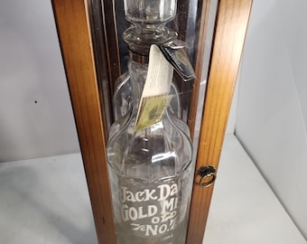 Puede incluir: Una jarra de cristal transparente con tapón, dentro de una vitrina de madera. La jarra tiene las palabras "Jack Daniel's Gold Medal Old No. 7" grabadas en la parte delantera. La vitrina de madera tiene una parte delantera de cristal y un pequeño pestillo color latón.