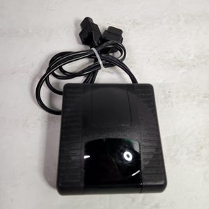 May include: Black rectangular electronic device with a black cord and two black plugs. The device has a black, rectangular button on the front.
