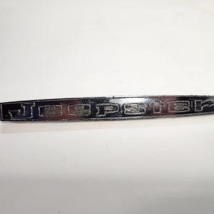 May include: A silver metal emblem with the word "Jeepster" engraved in black lettering.
