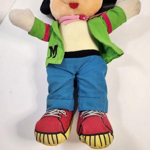 May include: A plush Mickey Mouse toy wearing a green jacket with a pink bandana, blue jeans, and red trainers. The jacket has a white "M" on the front.