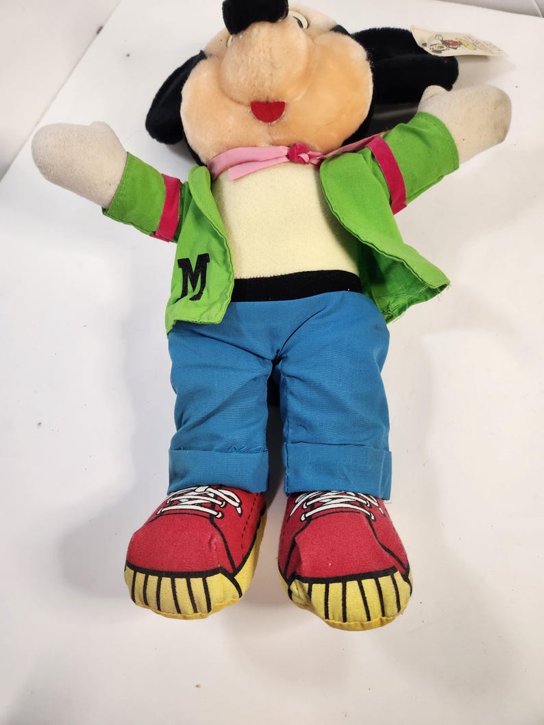 May include: A plush Mickey Mouse toy wearing a green jacket with a pink bandana, blue jeans, and red trainers. The jacket has a white "M" on the front.