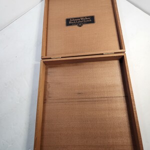 Puede incluir: Caja de madera abierta con tapa con bisagras. La caja es de color marr&oacute;n claro y tiene forma rectangular. La tapa tiene una peque&ntilde;a etiqueta negra rectangular con las palabras "Johnnie Walker Black Label Scotch 12 Years Old."