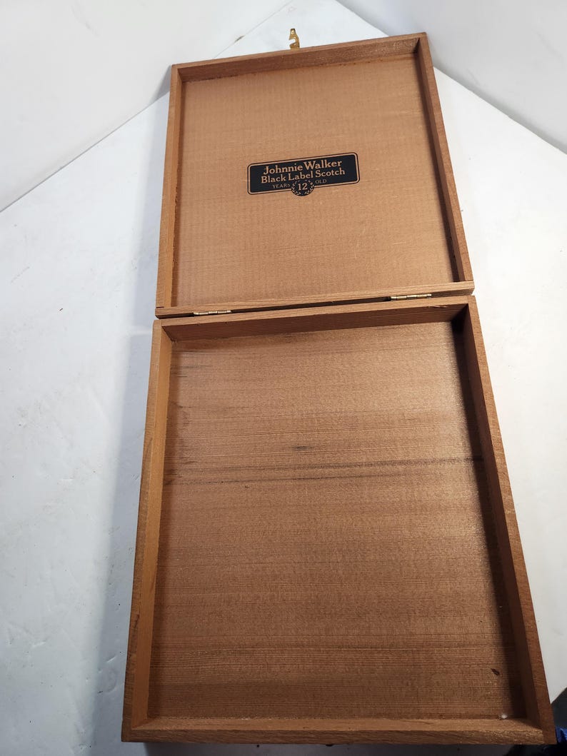 Puede incluir: Caja de madera abierta con tapa con bisagras. La caja es de color marr&oacute;n claro y tiene forma rectangular. La tapa tiene una peque&ntilde;a etiqueta negra rectangular con las palabras "Johnnie Walker Black Label Scotch 12 Years Old."