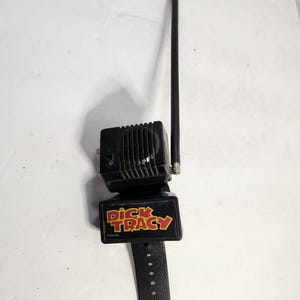 May include: A black Dick Tracy wrist radio toy with a black antenna, speaker, and watch band. The watch face has the Dick Tracy logo in red and yellow. The toy is a vintage collectible.