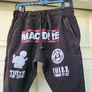 May include: Dark gray sweat shorts with white and red text that reads "MAC DERE" and "THIZZ OR DIE". The shorts have a drawstring and pockets. A white graphic of a person is on the left leg. The shorts are hanging on a black hanger.