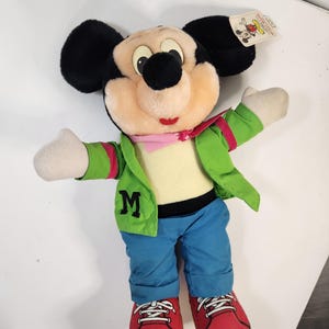 May include: A plush Mickey Mouse toy wearing a green jacket with a red stripe, blue jeans, and red shoes. The jacket has a large "M" on the front.