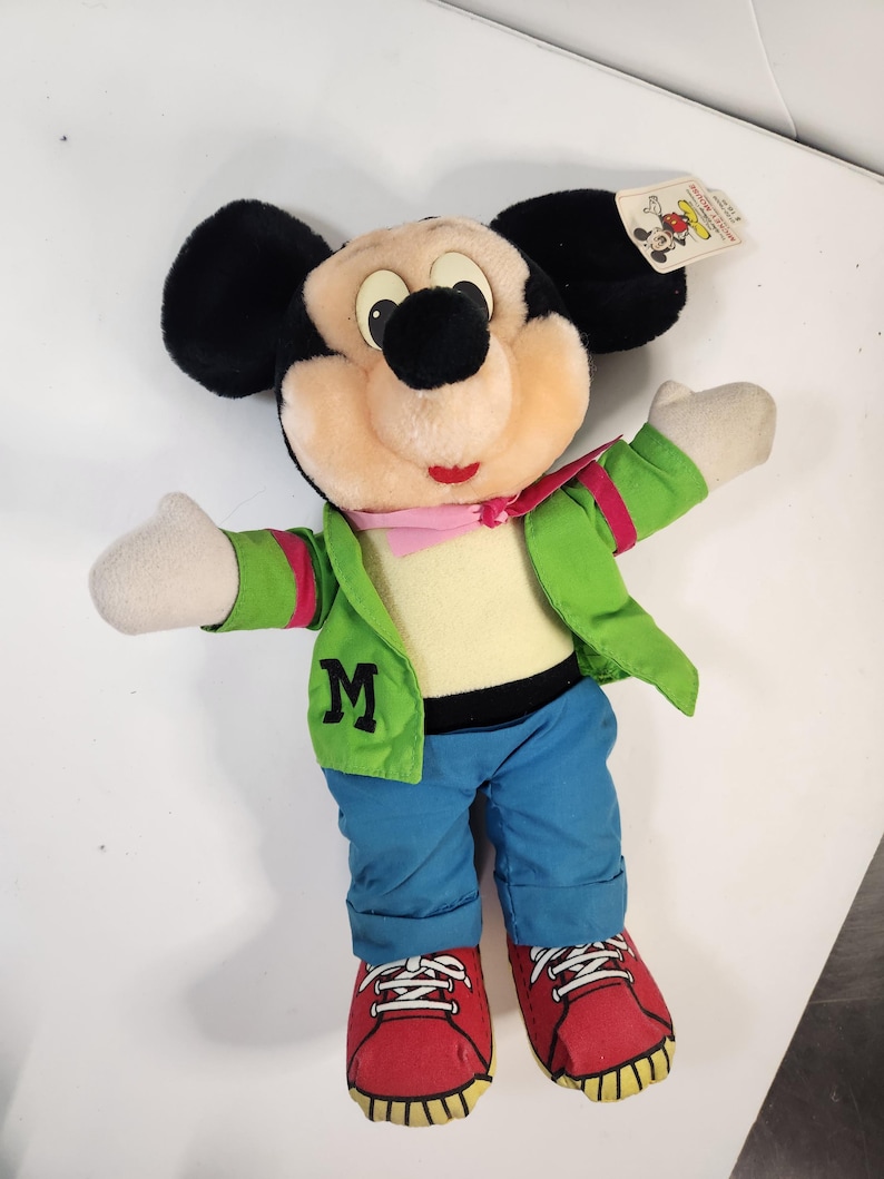 May include: A plush Mickey Mouse toy wearing a green jacket with a red stripe, blue jeans, and red shoes. The jacket has a large "M" on the front.
