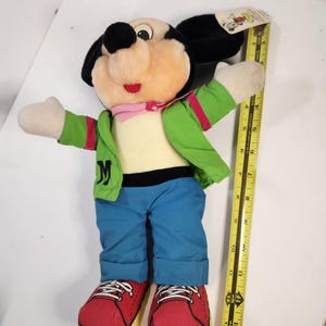 May include: A plush Mickey Mouse toy wearing a green jacket, blue jeans, and red trainers. The toy is approximately 30 cm tall.