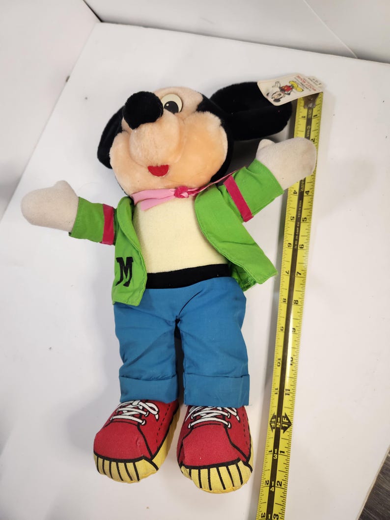 May include: A plush Mickey Mouse toy wearing a green jacket, blue jeans, and red trainers. The toy is approximately 30 cm tall.