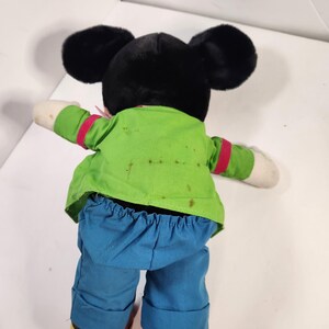 May include: A plush Mickey Mouse toy wearing a green shirt, red tie, and blue trousers. The toy has black fur and large ears.