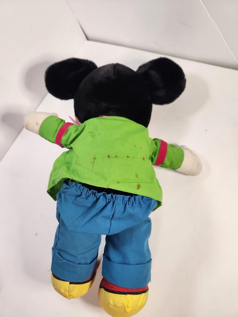 May include: A plush Mickey Mouse toy wearing a green shirt, red tie, and blue trousers. The toy has black fur and large ears.