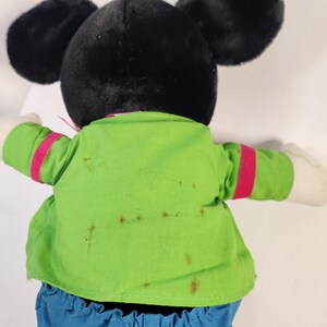 May include: A black and green plush Mickey Mouse toy wearing a green shirt with red trim and blue trousers. The toy has brown stains on the shirt.