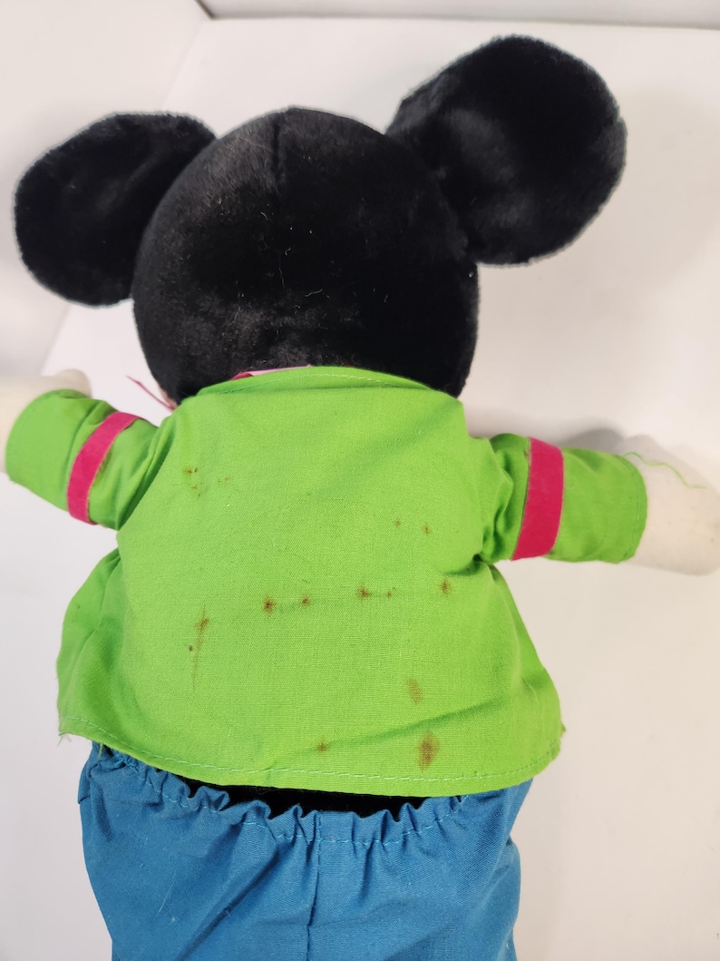 May include: A black and green plush Mickey Mouse toy wearing a green shirt with red trim and blue trousers. The toy has brown stains on the shirt.