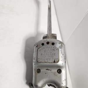 May include: A silver Signal-Stat 900 Sigflare with a green and red light, a lever, and the text "SIGNAL-STAT 900 SIGFLARE PAT. PEND. SIGNAL-STAT CORP. OKLYN, N.Y. U.S.A."