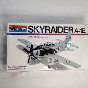 May include: A Monogram Skyraider A-1E model kit. The box features a detailed illustration of the aircraft in gray and black, with the text "The 'Flying Dump Truck' 1/72 Scale Model Kit". The box has red, white, and blue accents.