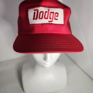 May include: A bright red baseball cap featuring a rectangular white patch on the front panel. The word Dodge is embroidered in red lettering across the center of the white patch. The cap has a structured crown and a flat brim with stitching.