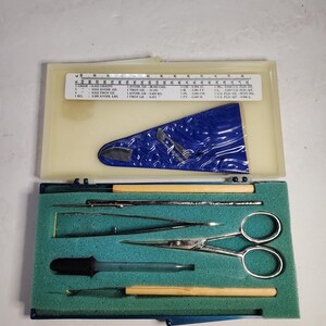 May include: An open, vintage medical instrument kit. The kit includes various stainless steel tools, such as scissors, tweezers, and probes, neatly arranged on a green foam insert. The lid features a ruler and a blue, plastic insert with text.