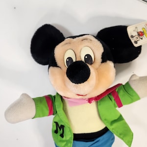 May include: A plush Mickey Mouse toy wearing a green jacket with a pink scarf, blue jeans, and a white shirt. The toy has a large 'M' on the jacket.