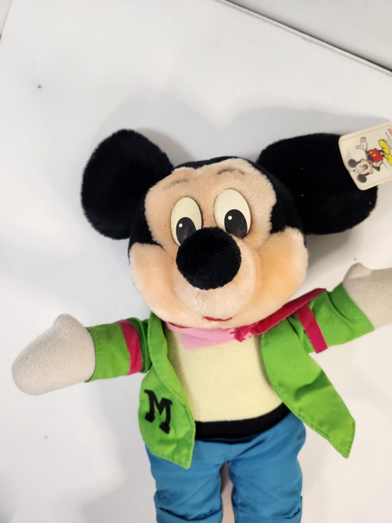 May include: A plush Mickey Mouse toy wearing a green jacket with a pink scarf, blue jeans, and a white shirt. The toy has a large 'M' on the jacket.