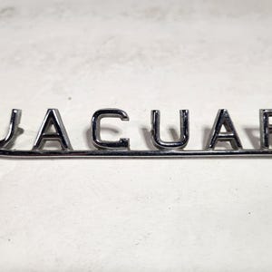 May include: A silver metal Jaguar emblem with the word "JAGUAR" spelled out in individual letters.