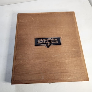Puede incluir: Una caja de madera rectangular de color marr&oacute;n claro. La superficie es texturizada. Una etiqueta negra y dorada en la parte delantera dice "Johnnie Walker Black Label Scotch YEARS 12 OLD".