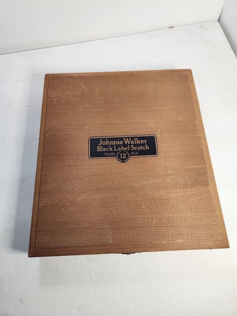 Puede incluir: Una caja de madera rectangular de color marr&oacute;n claro. La superficie es texturizada. Una etiqueta negra y dorada en la parte delantera dice "Johnnie Walker Black Label Scotch YEARS 12 OLD".