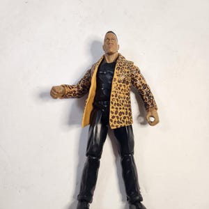 May include: Action figure of a man wearing a black shirt, black pants, and black shoes. The figure is wearing a tan and brown leopard print jacket. The figure is standing with arms slightly outstretched.