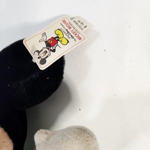 May include: A black and white plush Mickey Mouse toy with a green shirt and red shorts. The tag attached to the toy reads "Mickey Mouse" and "The Walt Disney Company". The price tag reads "&pound;1.98".