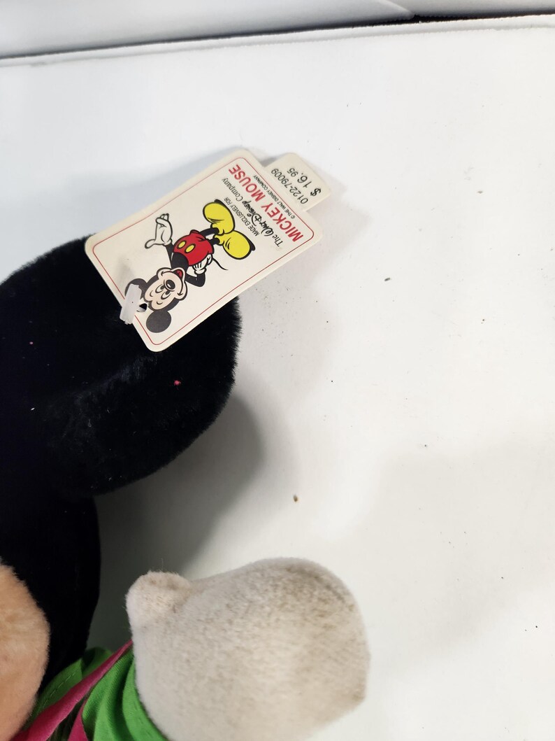 May include: A black and white plush Mickey Mouse toy with a green shirt and red shorts. The tag attached to the toy reads "Mickey Mouse" and "The Walt Disney Company". The price tag reads "&pound;1.98".