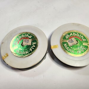 May include: Two white spools of "Cladding Mark Five" fiber-lubricated rolling line. Each spool has a green and gold label with text and a price tag. The line is black and wound around the spool.