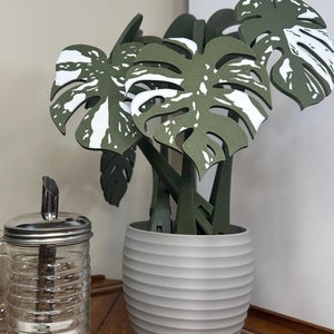 May include: A white ceramic planter with a ribbed texture holds a faux green and white monstera plant. The planter is on a wooden table with a sugar dispenser and a glass French press.