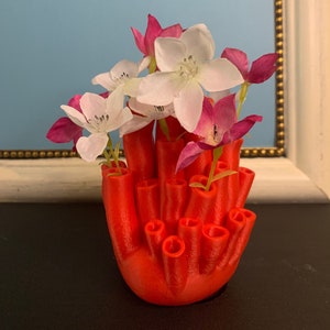 Könnte beinhalten: Eine rote, 3D-gedruckte Vase in Form eines Korallenriffs mit mehreren Öffnungen für Blumen. Die Vase ist mit weißen und rosa Blumen gefüllt.