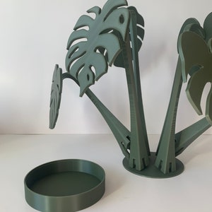 May include: A green, 3D printed planter with a round base and four stems that hold monstera leaf shapes. The planter is separated from the base, which is also green.