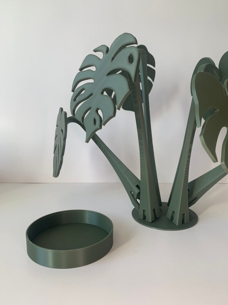 May include: A green, 3D printed planter with a round base and four stems that hold monstera leaf shapes. The planter is separated from the base, which is also green.