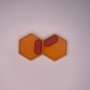 Może przedstawiać: Pomarańczowy sześciokątny organizer na tabletki z dwoma przegródkami, każda z czerwoną tabletką.