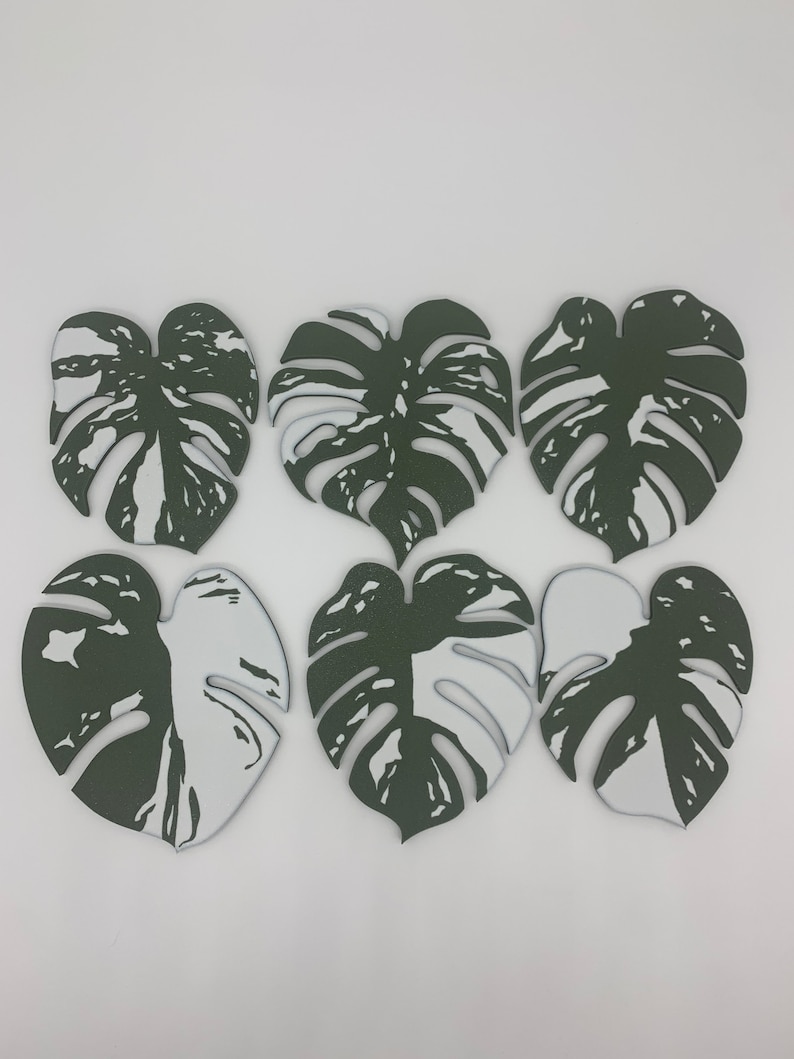 May include: Six green and white monstera leaf-shaped coasters. The coasters are made of a thick, durable material and have a unique, modern design.