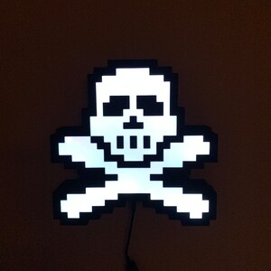 Könnte beinhalten: Ein weißes, pixeliges Totenkopf- und Knochenkreuz-Design, beleuchtet mit weißem Licht. Das Design ist an einem schwarzen Kabel befestigt.