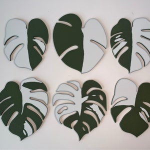 May include: Six green and white monstera leaf-shaped coasters. The coasters are painted half green and half white.