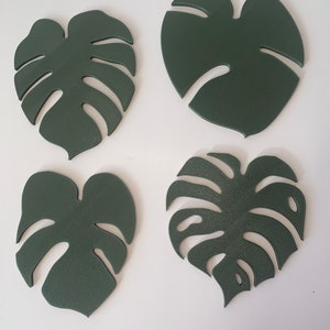 May include: Four green monstera leaf-shaped coasters. The coasters are made of a textured material and have a slightly raised design.