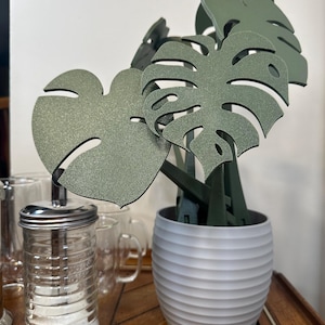 May include: A set of four green monstera leaf-shaped kitchen utensils in a gray planter with a ribbed texture. The utensils are made of a textured material and are arranged in a bouquet-like shape.
