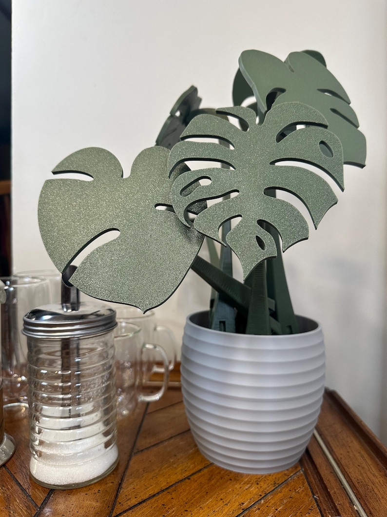 May include: A set of four green monstera leaf-shaped kitchen utensils in a gray planter with a ribbed texture. The utensils are made of a textured material and are arranged in a bouquet-like shape.