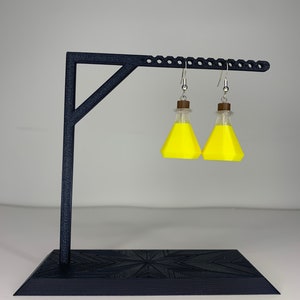 May include: A pair of yellow potion bottle earrings hanging from a black earring stand. The earrings are made of polymer clay and have a silver hook.