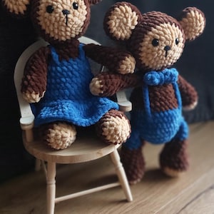 Puede incluir: Dos muñecos de mono de ganchillo hechos a mano. Uno está sentado en una pequeña silla de madera y lleva un vestido y un lazo azules. El otro mono lleva un peto azul y una pajarita. Ambos son marrones y beige.
